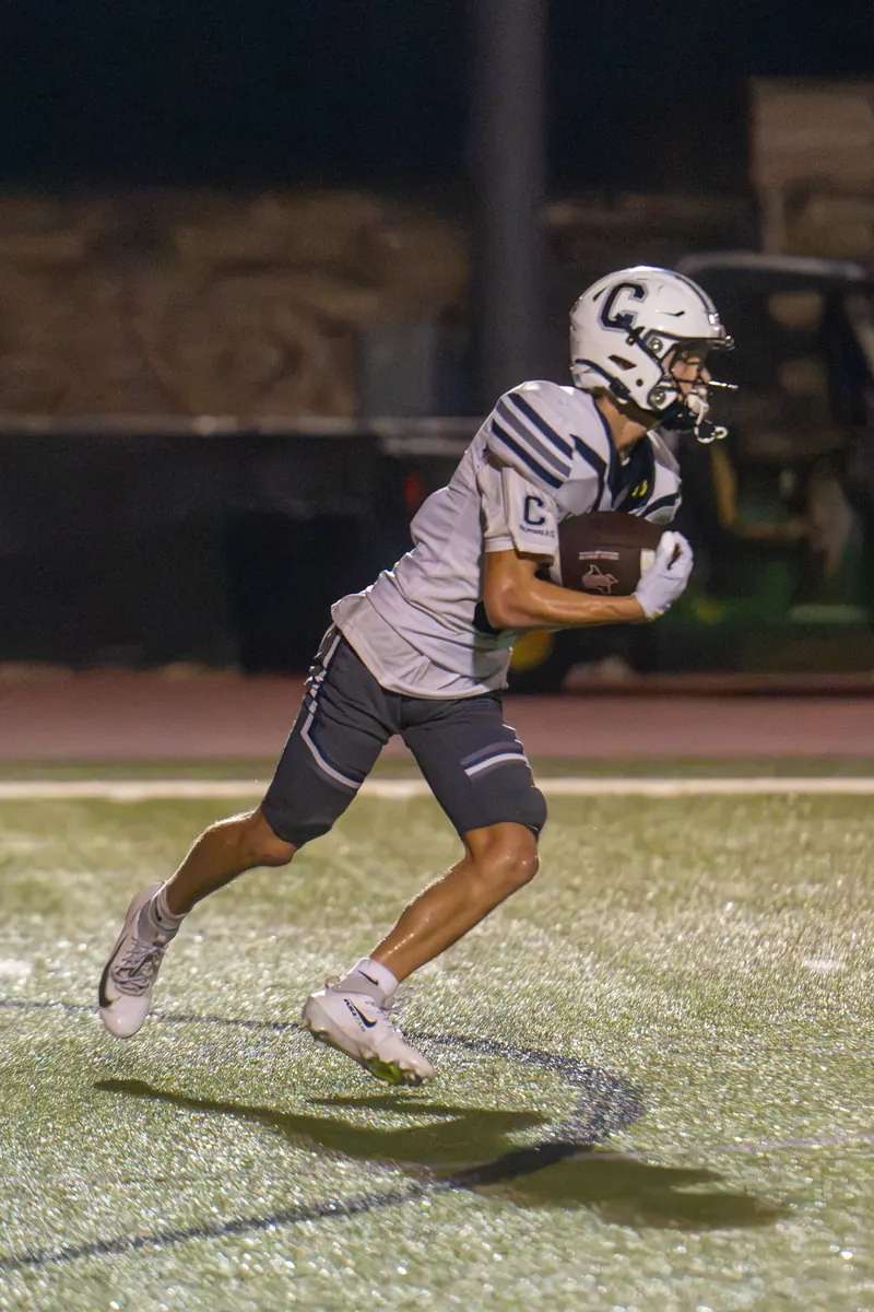 A football receiver in a gray road uniform tucks the ball and cuts upfield on a night game
