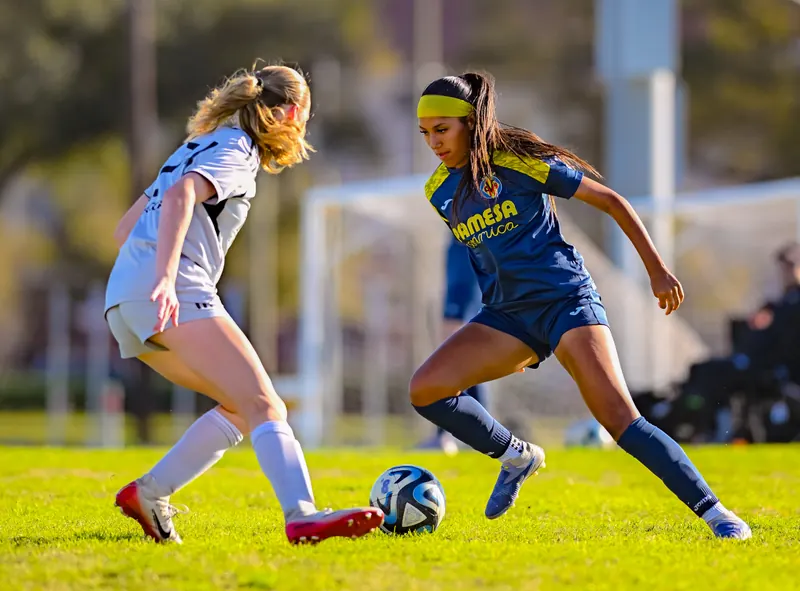 A girls club soccer player in a blue and yellow kit dribbles the ball past a defender on a grass field